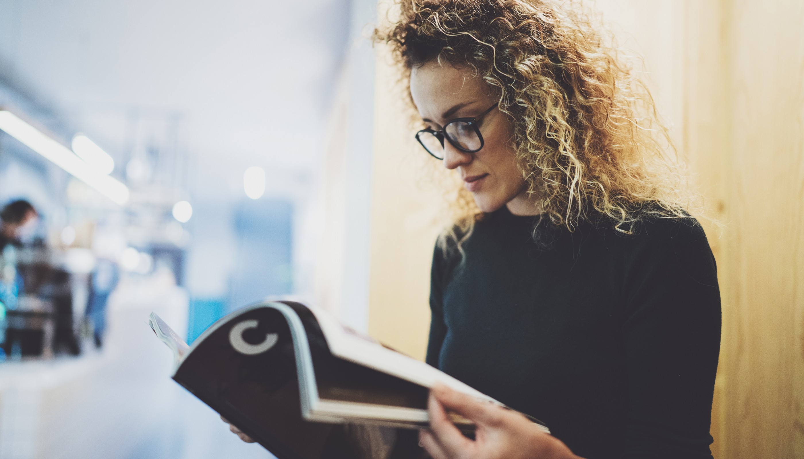 Charming fashionable woman with eyes glasses reading magazine sitting indoor in urban cafe.Casual portrait of pretty girl. Blurred background